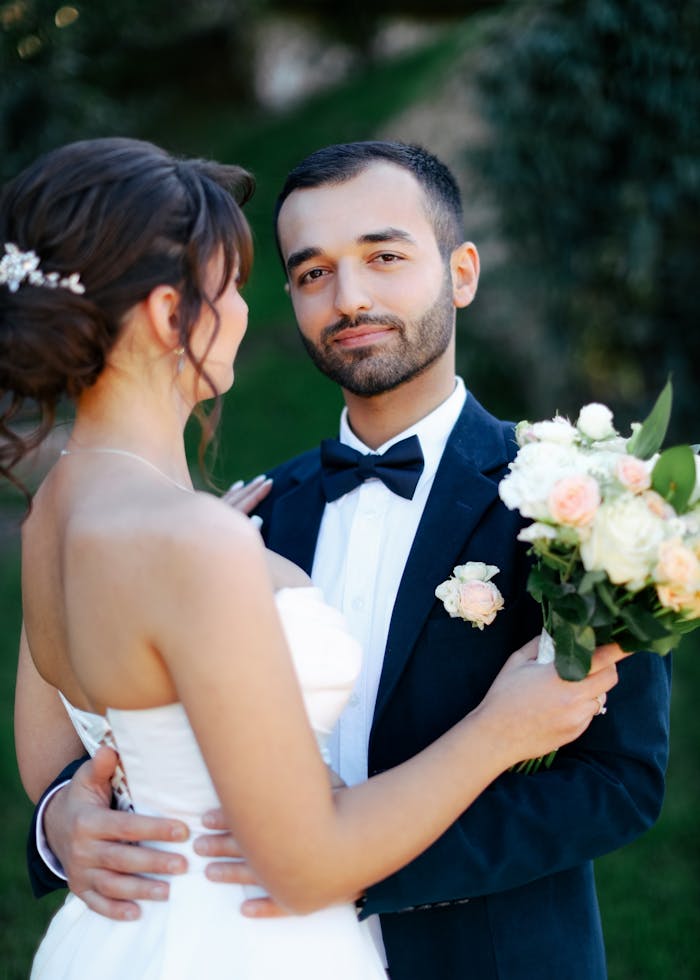 A loving couple in wedding attire sharing a moment outdoors with a bouquet.