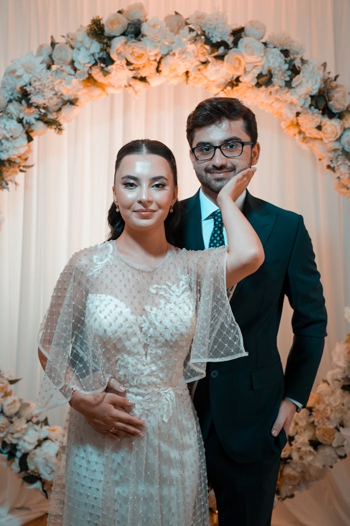 Charming bride and groom pose under a floral wedding arch indoors.