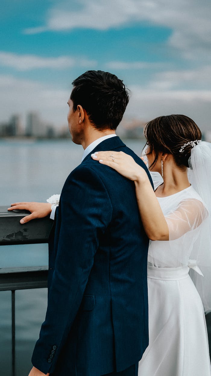 Bride and groom in elegant attire by the waterfront, sharing a peaceful moment.